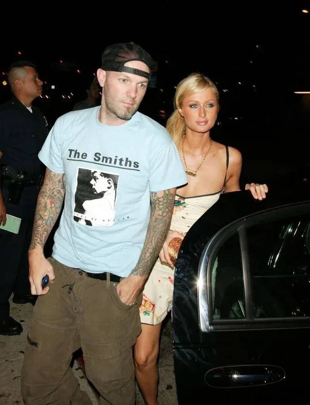 Man and Paris Hilton exiting a car. Man with tattoos wearing a Smiths t-shirt. Hilton in floral dress. Nighttime setting.