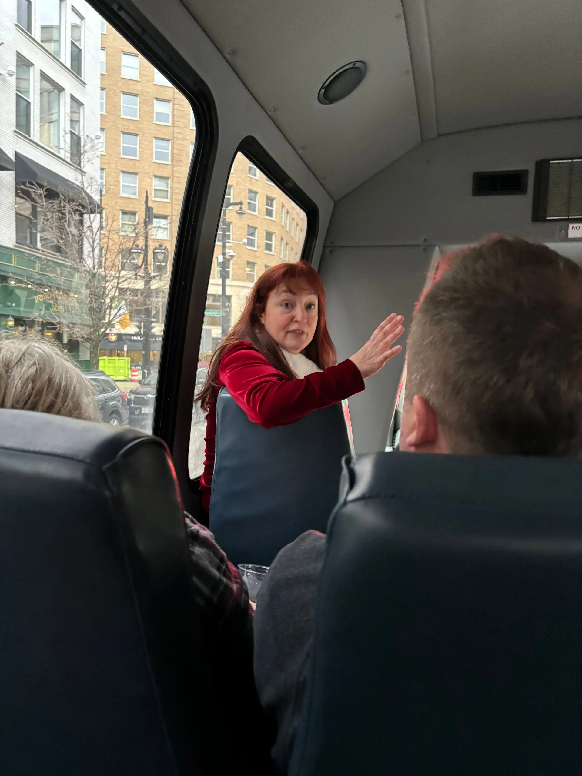 Woman in red jacket gestures while standing in bus aisle, city view outside window.