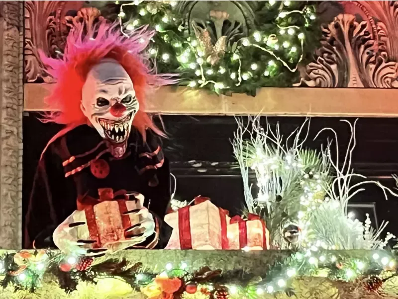 A clown is holding a gift box in front of a christmas tree.