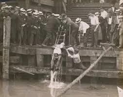 Men rescuing someone from water near a pier. Bystanders watch. Black and white photo.