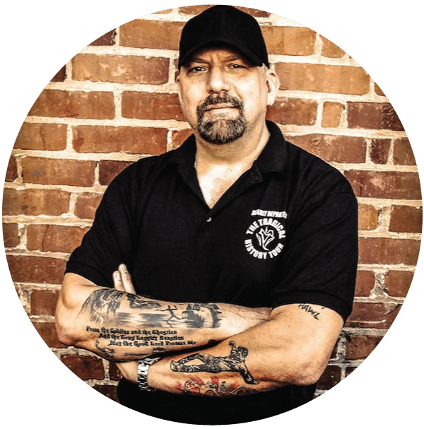 Man with tattoos and black cap, arms crossed, in front of a brick wall.