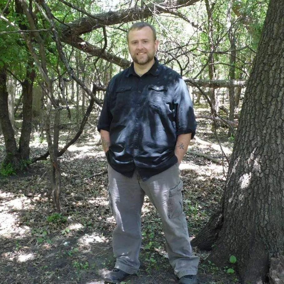 A man in a black shirt is standing in the woods with his hands in his pockets