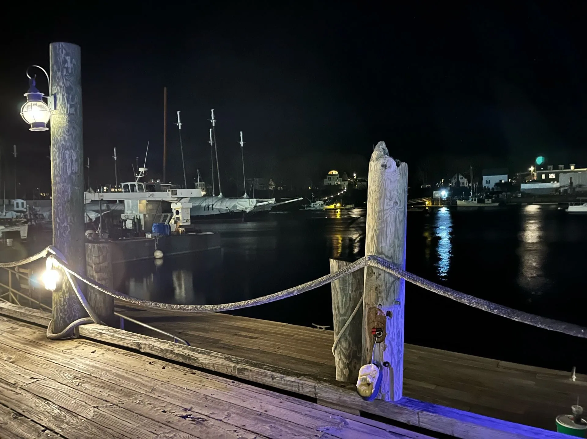 Night view of a wooden pier with rope railing, leading to a harbor with boats and illuminated buildings.