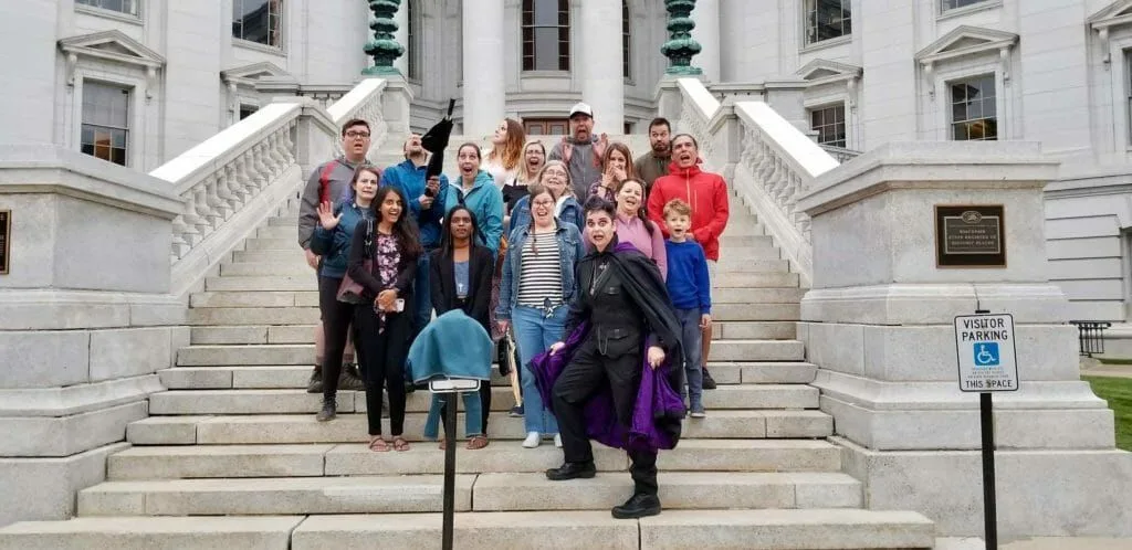 Group poses on stone steps in front of a white building with pillars. Some wave, smile, or look at the camera. One person holds a purple cloth.