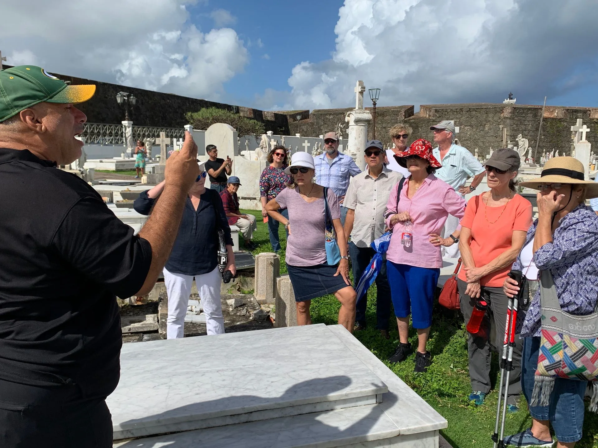Tour guide speaking to group in a cemetery. Several people look on, some wearing hats, on a sunny day.