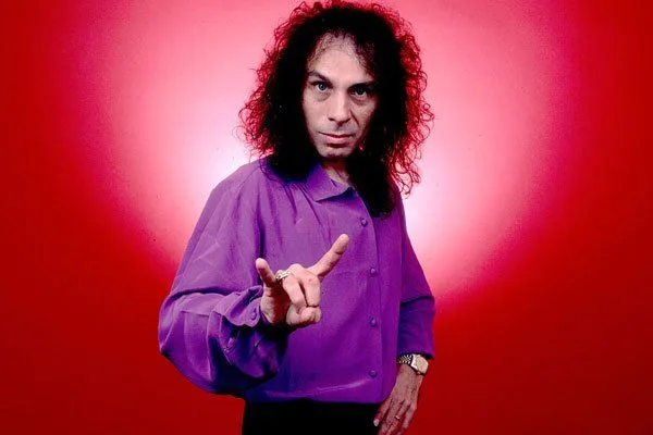 A man with long curly hair is wearing a purple shirt and pointing at the camera.