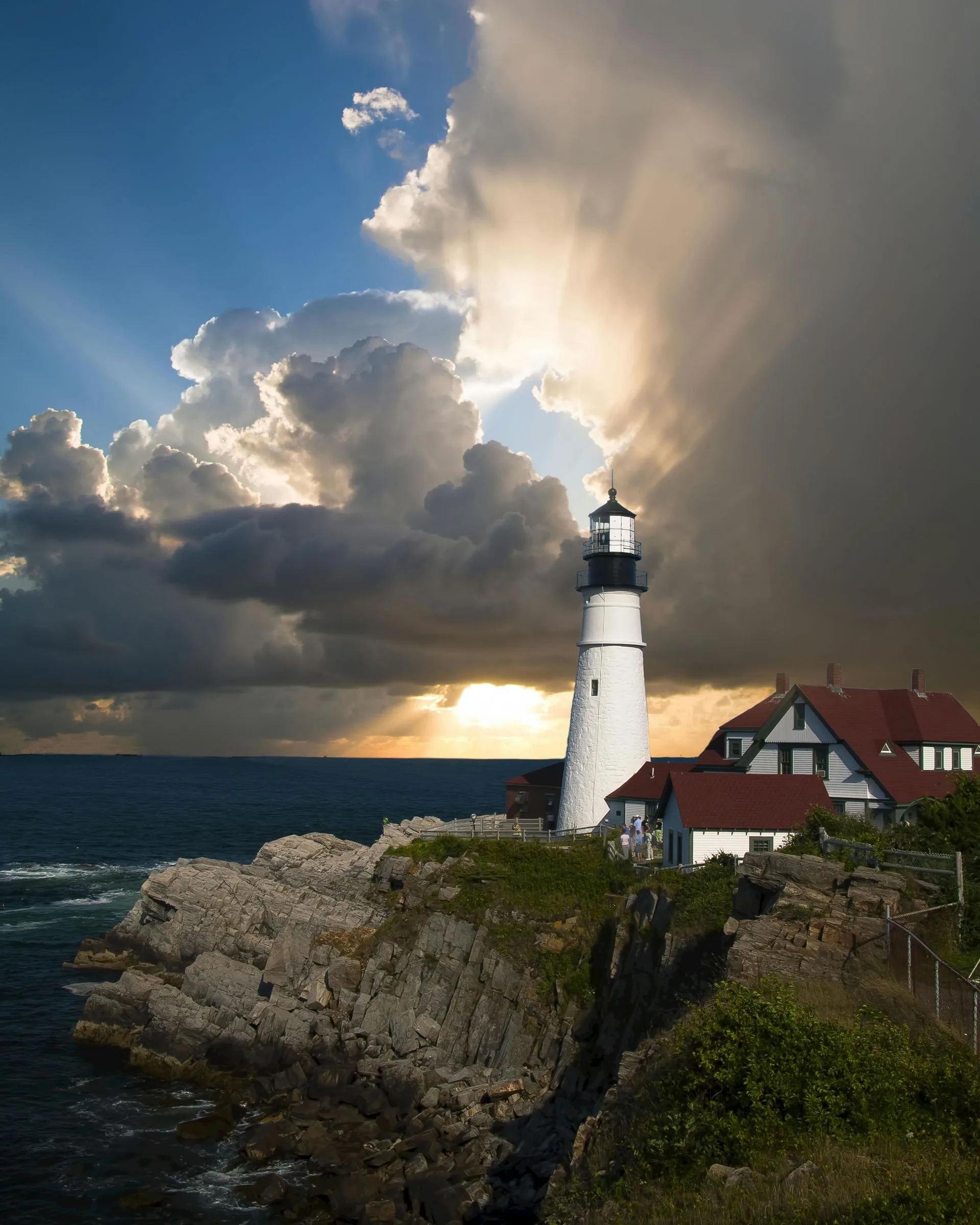 Lighthouse on rocky coast under dramatic cloudy sky, with sun rays breaking through.