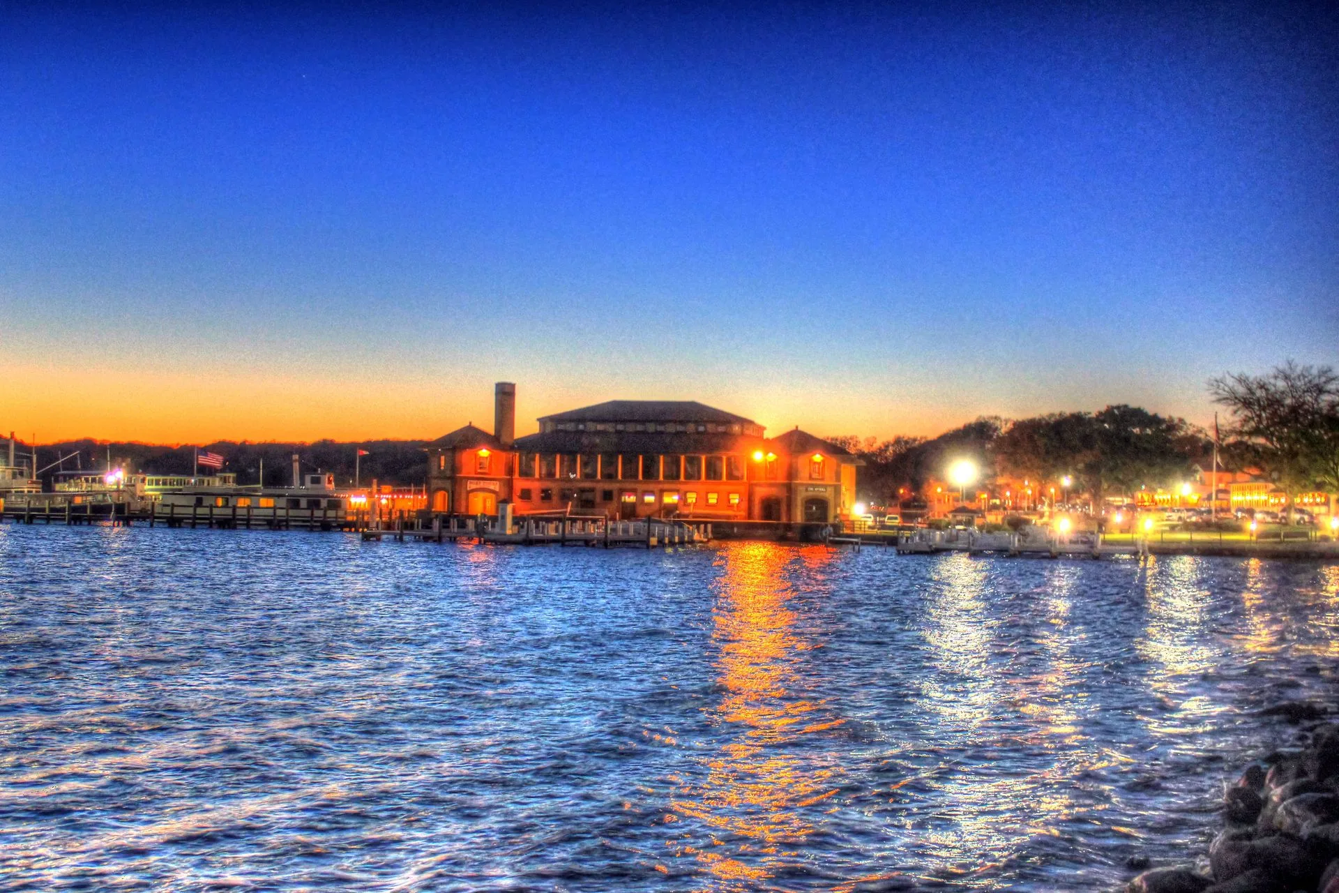 Waterfront building at dusk with illuminated lights reflected on the water under a blue sky.