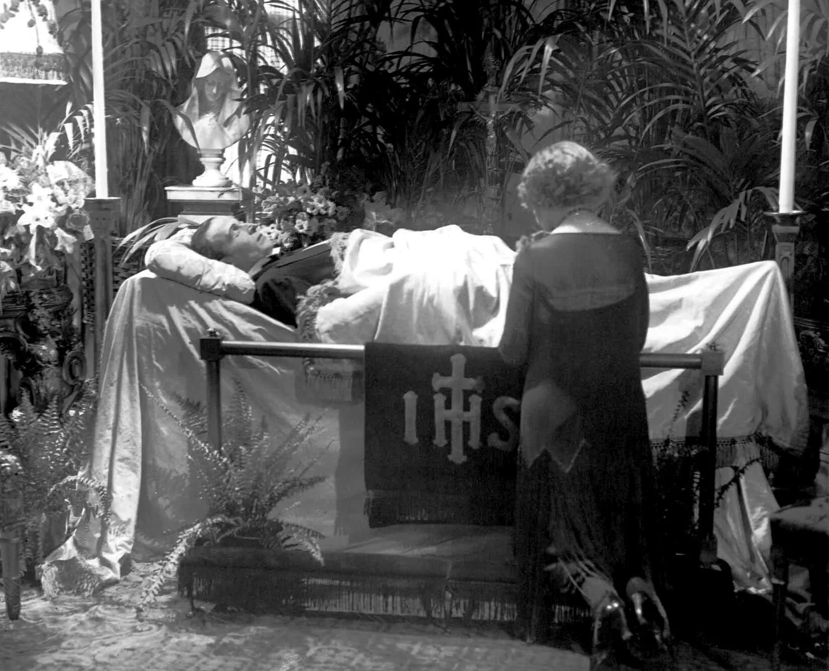 A woman is kneeling in front of a coffin with a banner that says ihs