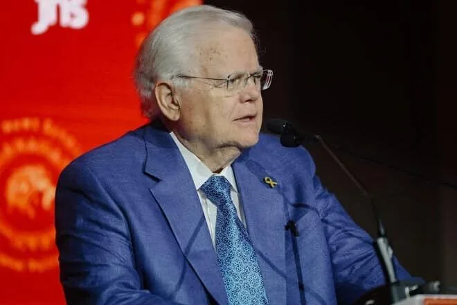 Man in blue suit speaking at a podium with a red background.
