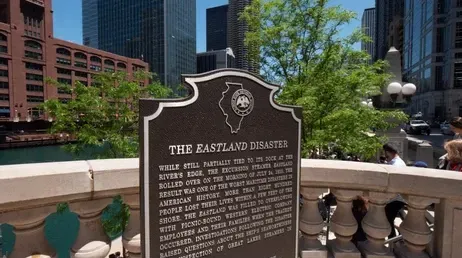 Historical marker for the Eastland Disaster in Chicago, near a river, with buildings and sky in the background.