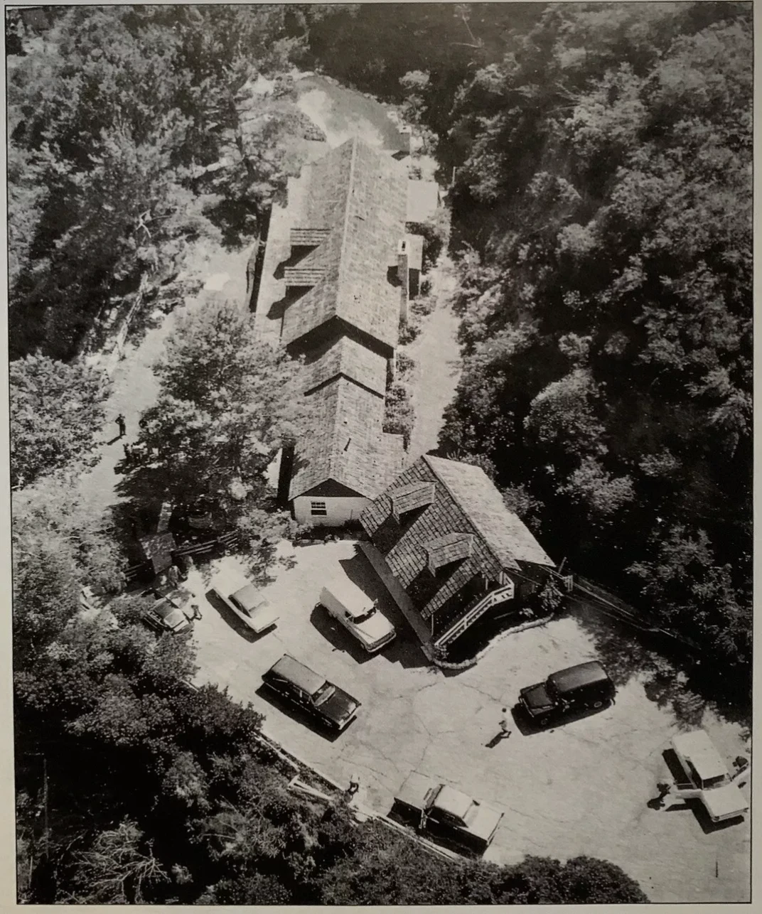 Aerial view of a long building nestled among trees, with cars parked out front. Black and white photo.