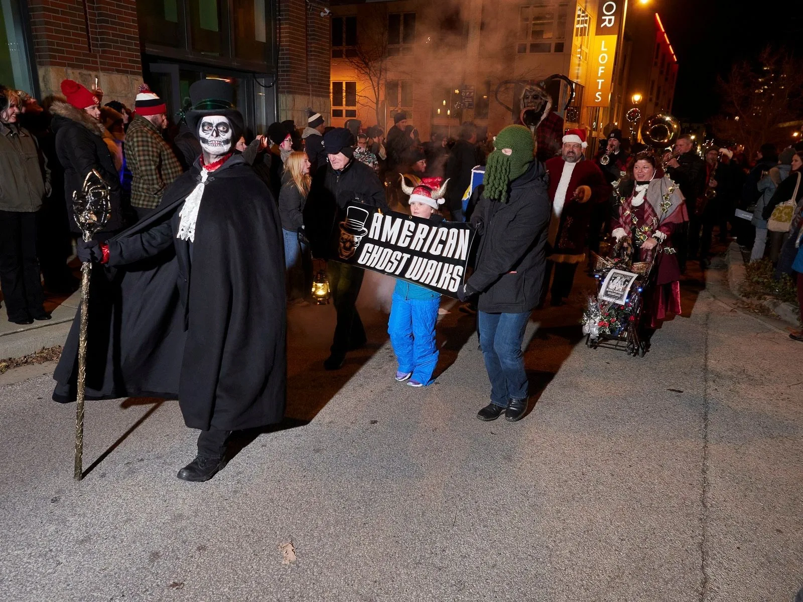 Street parade with costumed figures, including a person in a top hat and a child holding a sign that says 