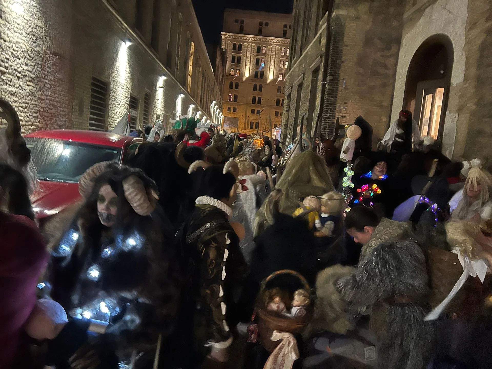 Costumed people crowd a street at night, illuminated by string lights. A red car is on the left.