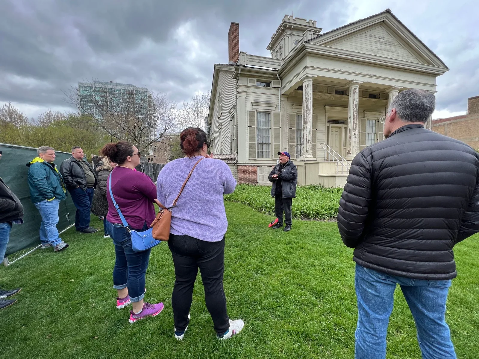Group of people on a tour in front of a white historic house with columns. Cloudy sky.