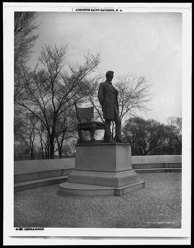 Statue of a man in a suit standing on a stone pedestal. Bare trees in the background.